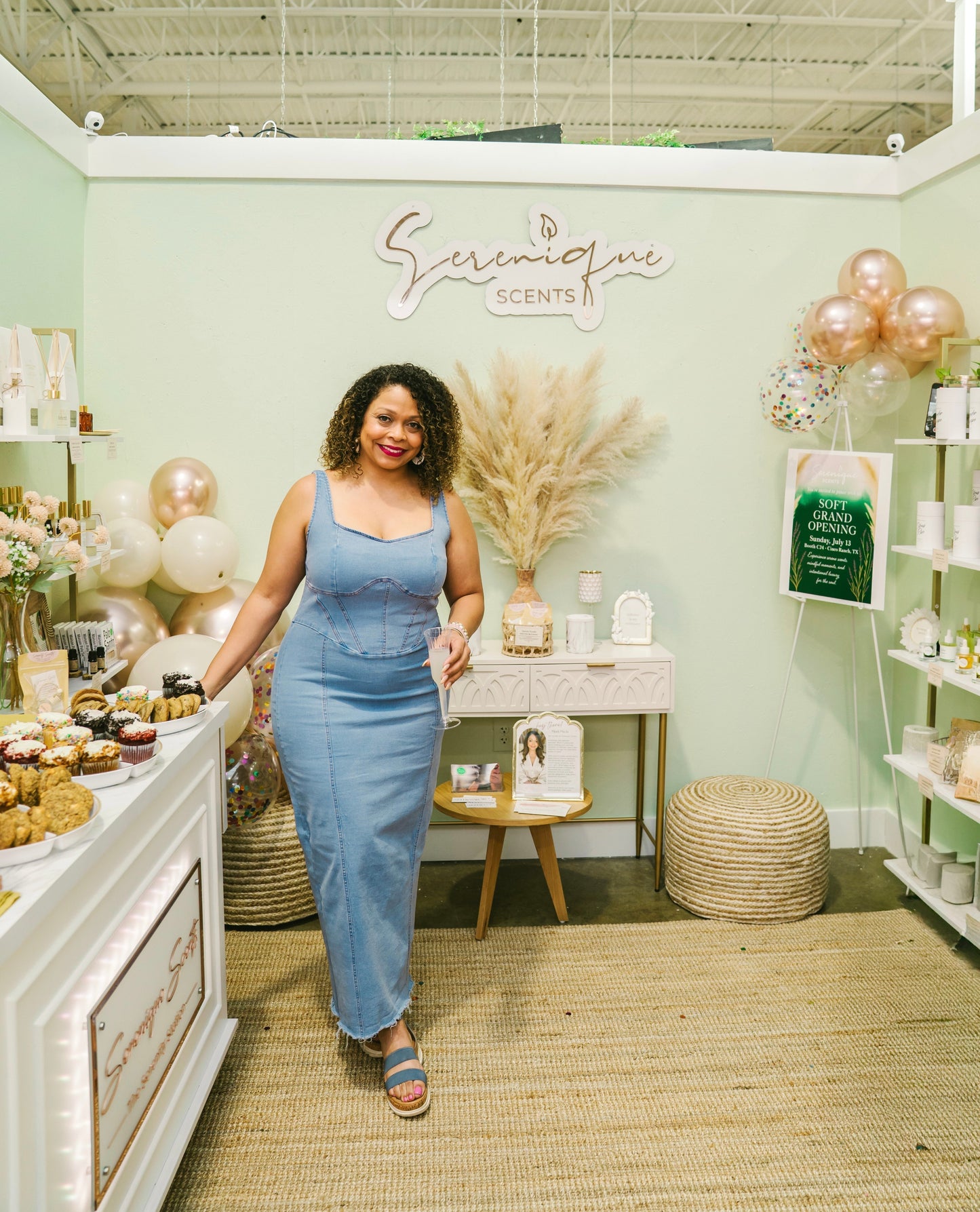 Michi Slaughter standing in front of the Serenique Scents booth at the Painted Tree Boutique, surrounded by product displays, balloons, and soft natural decor.